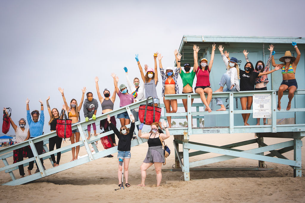 Women of the Yoniswell Surf Collective standing with their arms up in the air looking triumphant along a ramp leading up to a wooden beach hut, many are wearing masks during Covid